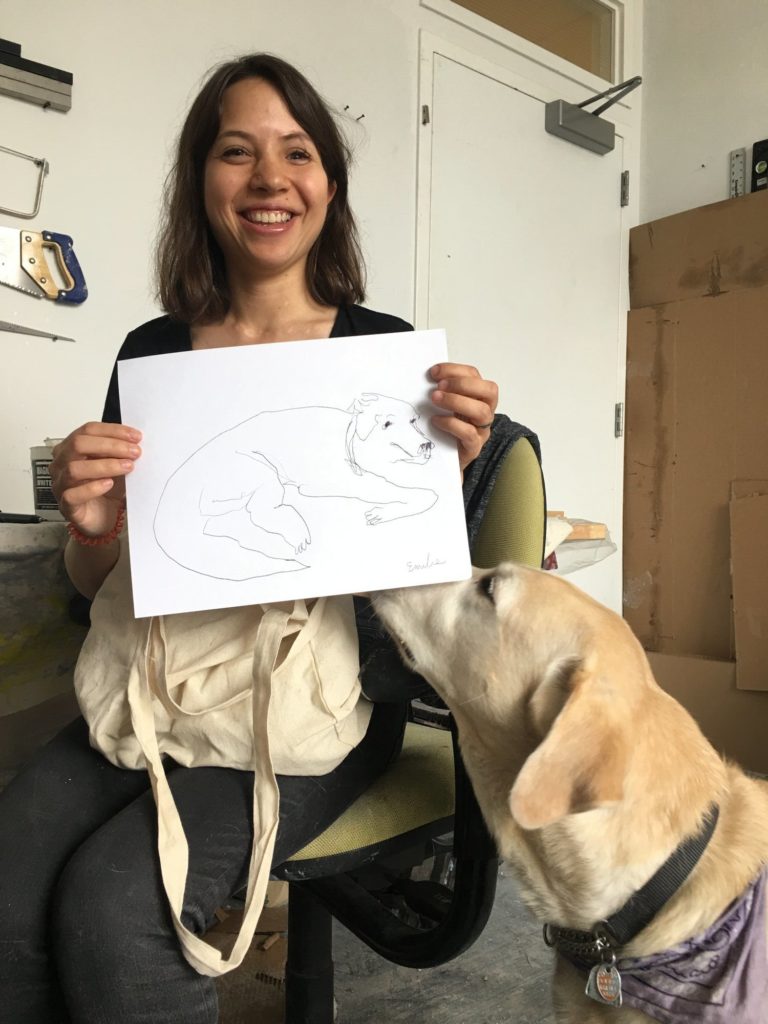 Artist Emilie Gossiaux is smiling holding a piece of paper with a line drawing of her guide dog London. London, a yellow Labrador Retriever is sniffing the piece of paper.