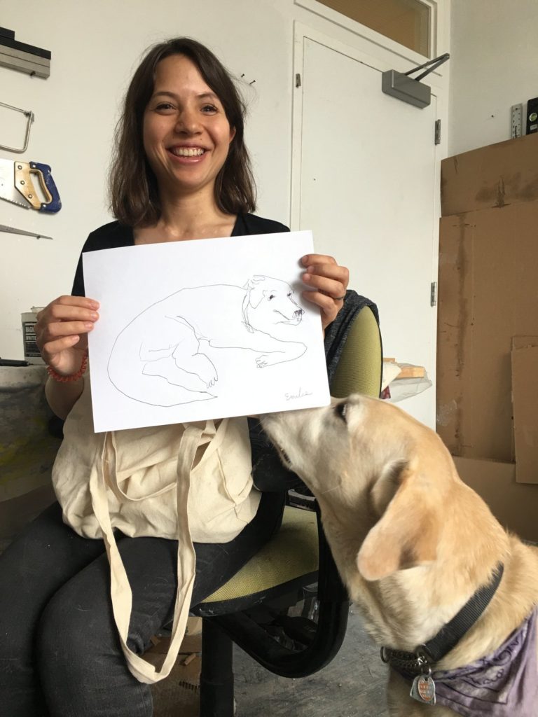 Artist Emilie Gossiaux is smiling holding a piece of paper with a line drawing of her guide dog London. London, a yellow Labrador Retriever is sniffing the piece of paper.